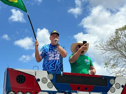 Coronel Hélio, de boné e camisa azul com o número 22, discursa ao microfone em cima de um trio e segura uma bandeira do Brasil