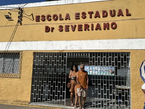 Casal sai pelo portão gradeado da Escola Estadual Dr. Severiano, de fachada amarela, enquanto uma mulher passa com guarda-chuva