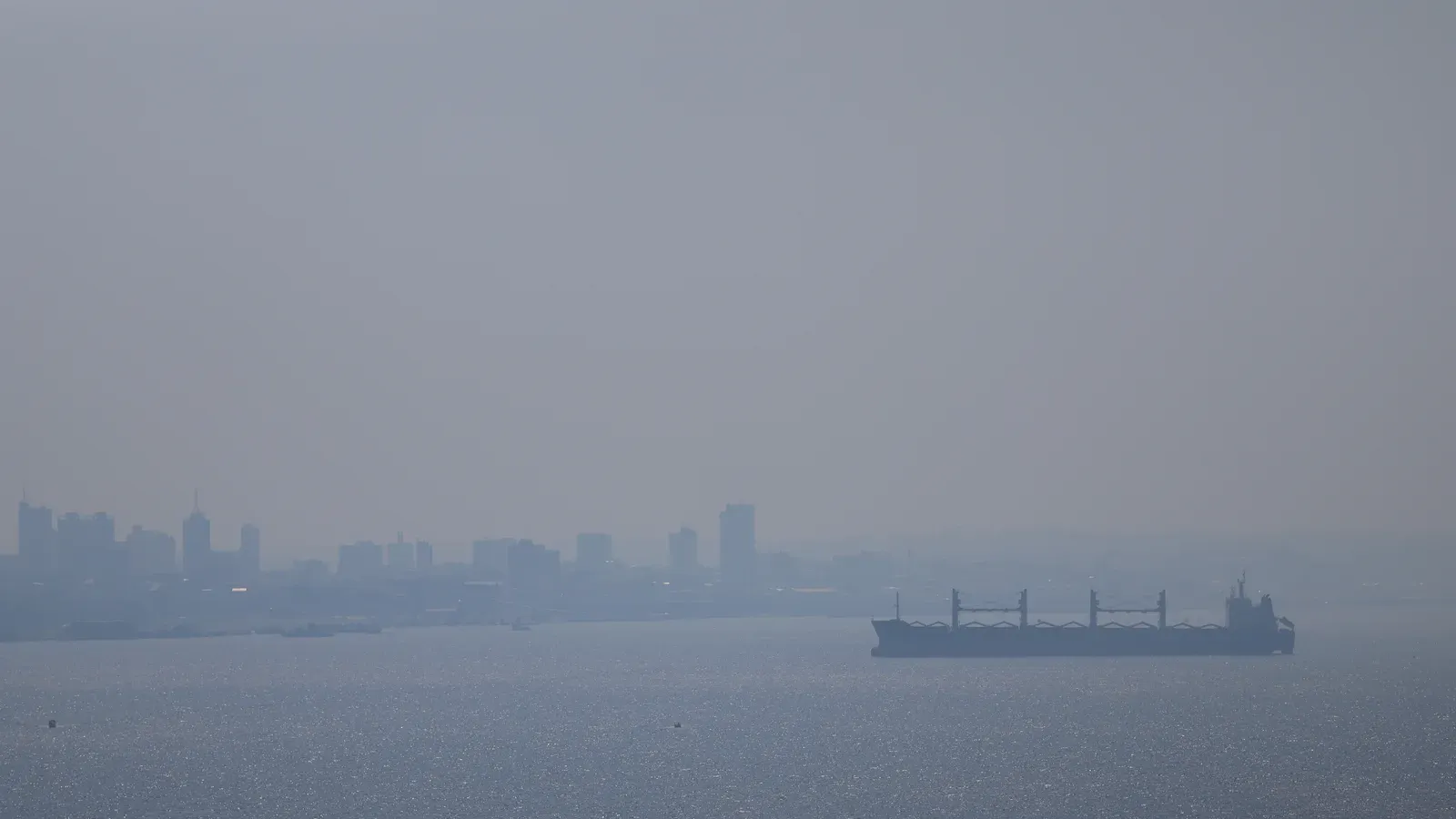 Prédios de Manaus quase sumindo na fumaça acinzentada, com um navio cargueiro no Rio Negro em primeiro plano