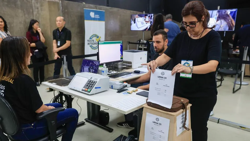 Servidora da Justiça Eleitoral segura cédula ao lado de urna eletrônica durante teste de integridade