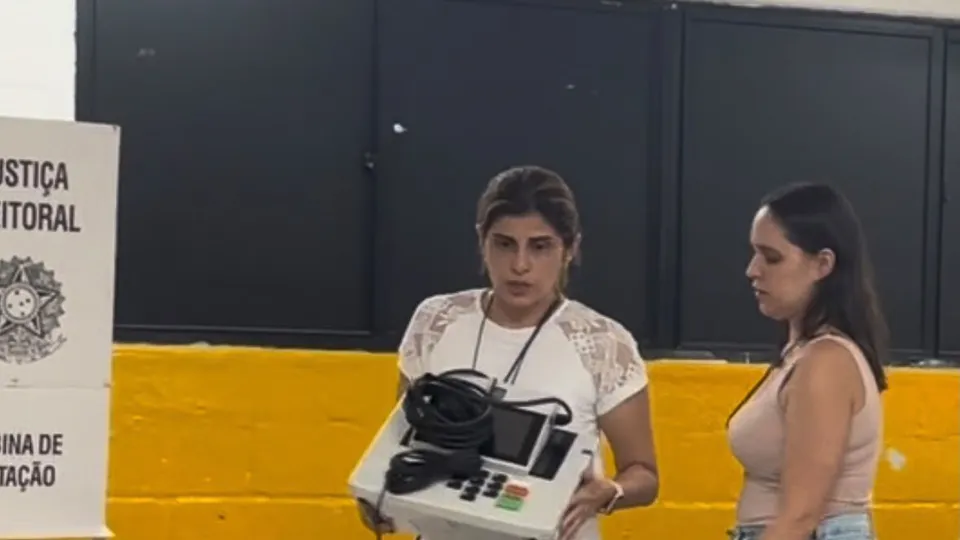 Mesária carrega a urna eletrônica com defeito ao lado da cabine de votação